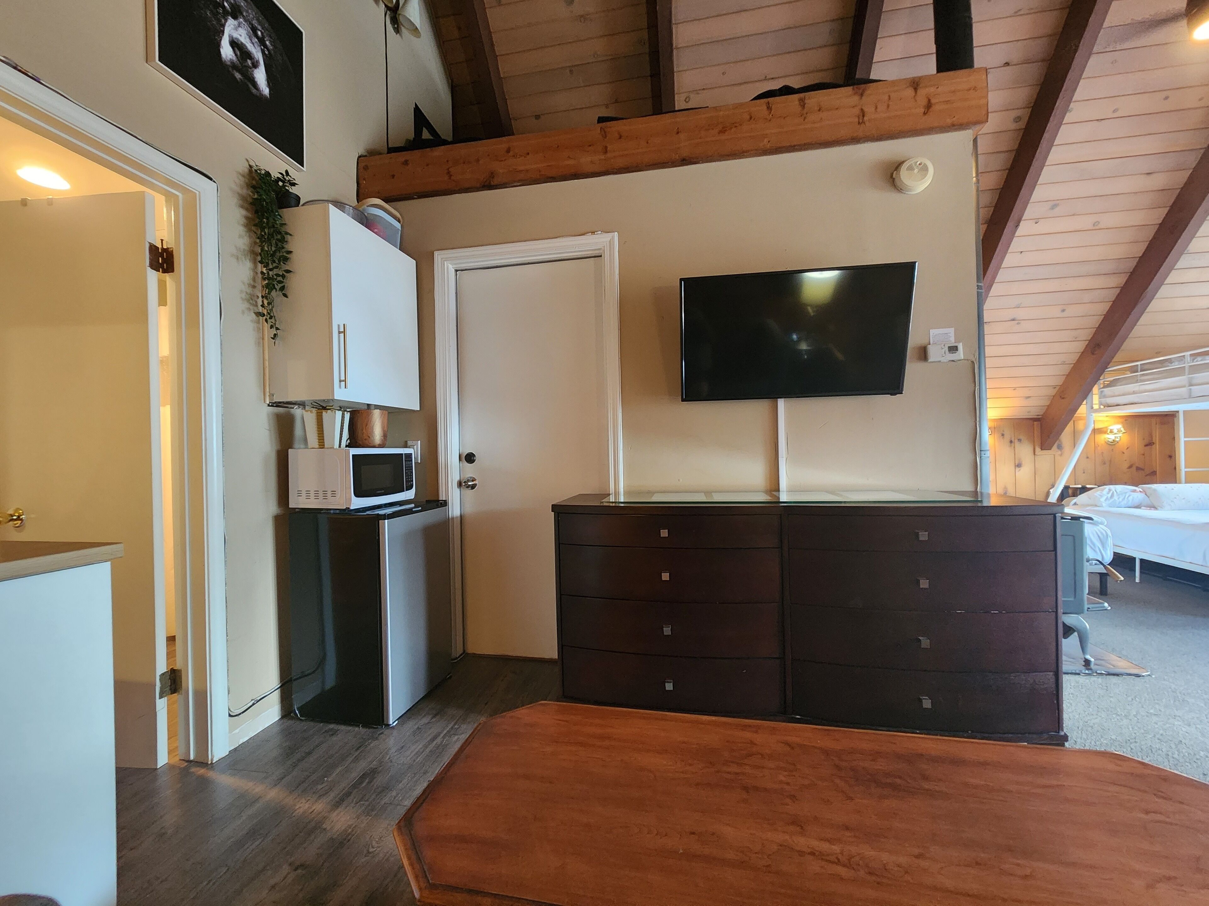 A compact room with a kitchenette, a flat-screen TV mounted on the wall, a wooden ceiling, and a bed with a bunk.