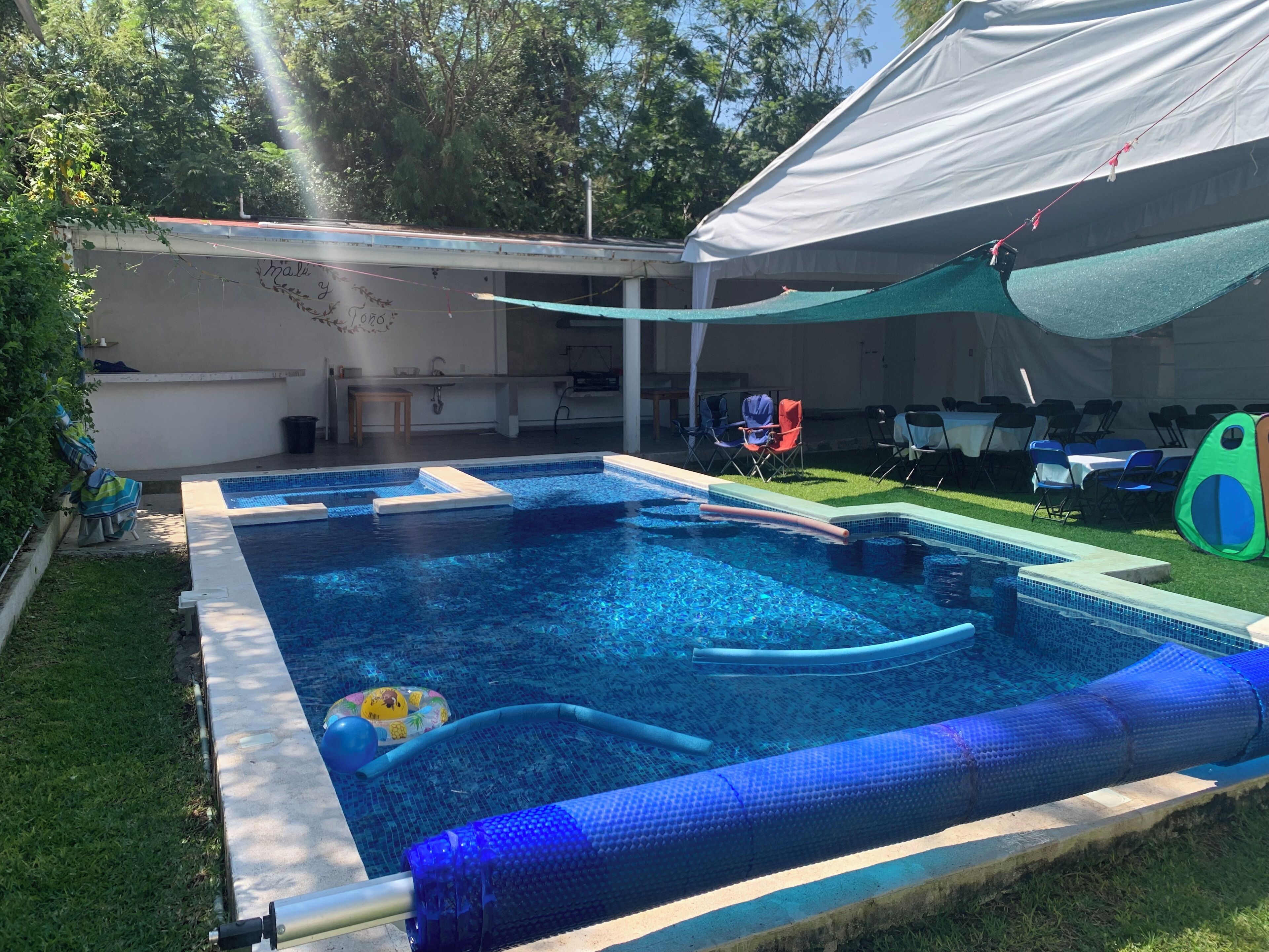 A swimming pool with a white tent overhead, surrounded by grass and trees.