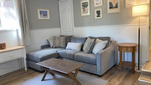 Un salon chaleureux avec un canapé d’angle gris, une table basse en bois et une petite table d’appoint ronde. Des tableaux encadrés ornent le mur, il y a une porte blanche et une fenêtre avec des rideaux.