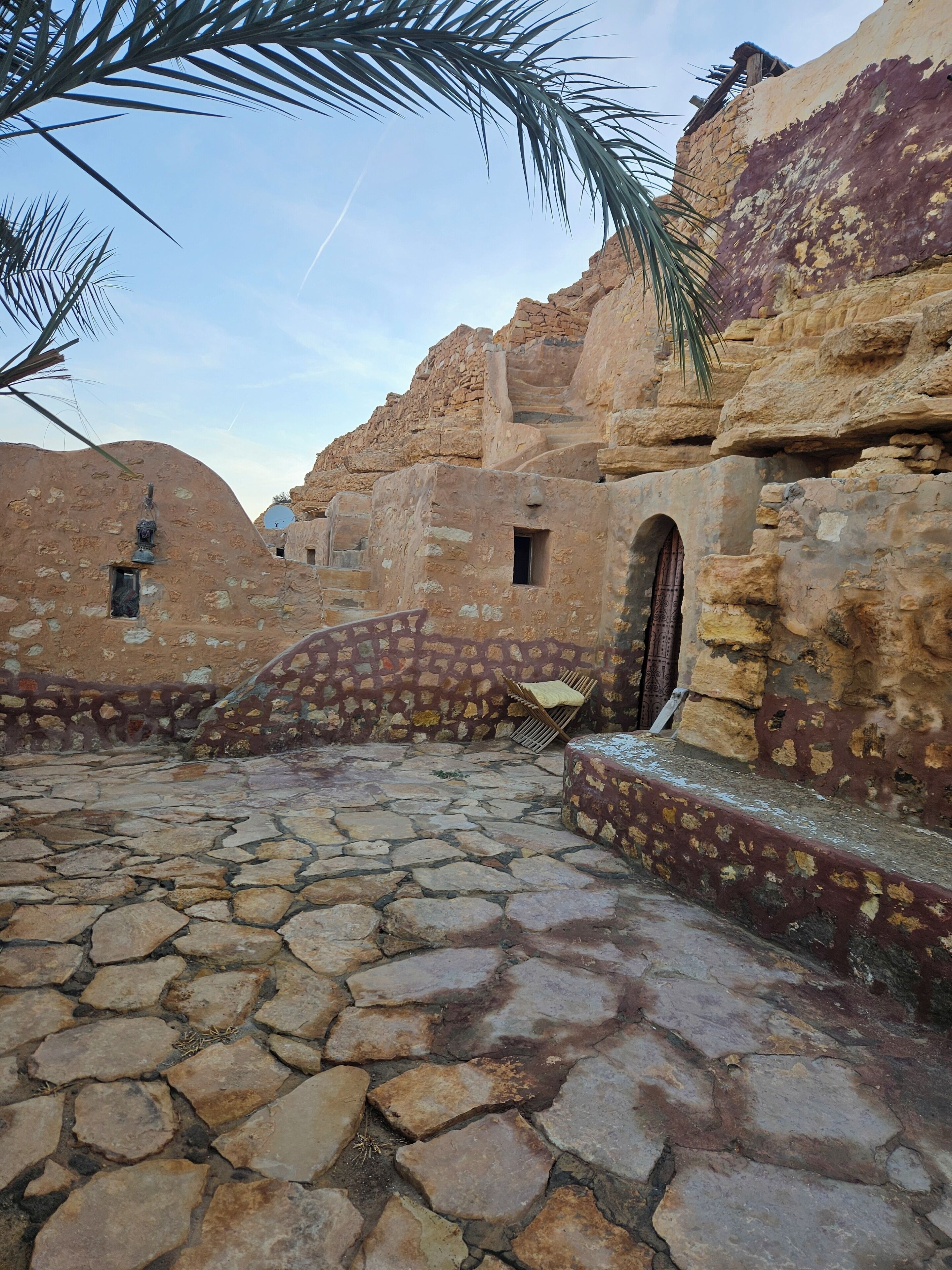 Un patio con suelo de piedra y arquitectura tradicional, que incluye un pequeño arco de entrada y un banco.