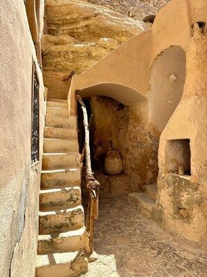 A narrow, stone-walled passageway with a wooden handrail and a small, arched doorway leading to a room with a tiled floor. - TROGLODYTIC RESIDENCE KENZA-CHENINI (Chenini)