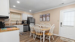 A compact kitchen with white cabinets, a wooden dining table with four chairs, a refrigerator, and a coffee maker.