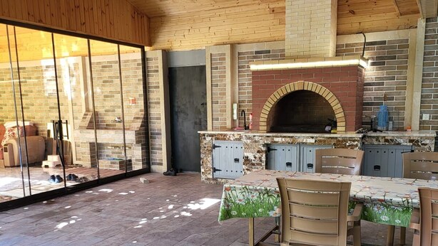Un espace cuisine extérieur doté d’un four en briques, d’une table à manger avec des chaises et d’une véranda donnant sur une terrasse.