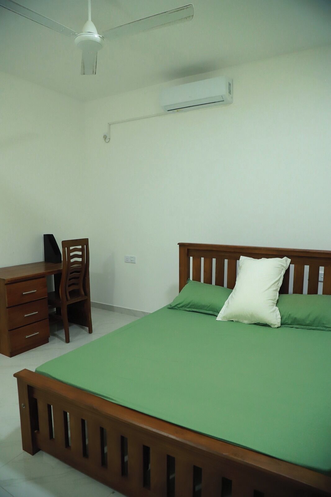 A bedroom with a wooden bed frame, green bedding, a desk with a chair, and an air conditioning unit on the wall.