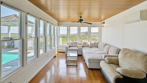 A spacious living room with a large sectional sofa, a glass coffee table, and a ceiling fan. Large windows offer views of a pool and outdoor area. - Waterfront Westhampton Haven: Private Beach (Westhampton Beach)