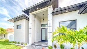 A modern two-story house with a covered entrance, a front door, and a small garden with plants and a pathway. - Charming 4-bedroom house with Heated Pool (Cape Coral)