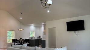 A spacious living room with a grey sectional sofa, a chandelier, a flat-screen TV mounted on the wall, and a kitchen in the background.