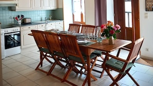 Un comedor con sillas de madera y una mesa preparada para cuatro, una cocina con armarios blancos y una estufa, y un techo de madera con vigas a la vista.