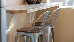 Un coin cuisine doté d’un plan de travail en marbre, de deux tabourets de bar en métal avec des assises en bois, et d’un saladier rempli de fruits posé sur le plan de travail.