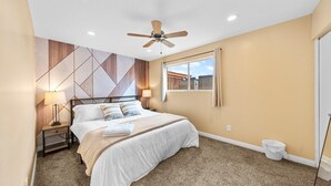 Une chambre à coucher comprenant un lit, une table de chevet, une lampe, un ventilateur de plafond, une fenêtre avec des rideaux et un mur orné de motifs géométriques.