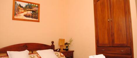 Un dormitorio con cabecera de madera, cama con ropa de cama estampada, mesita de noche con lámpara y florero, y un gran armario de madera.
