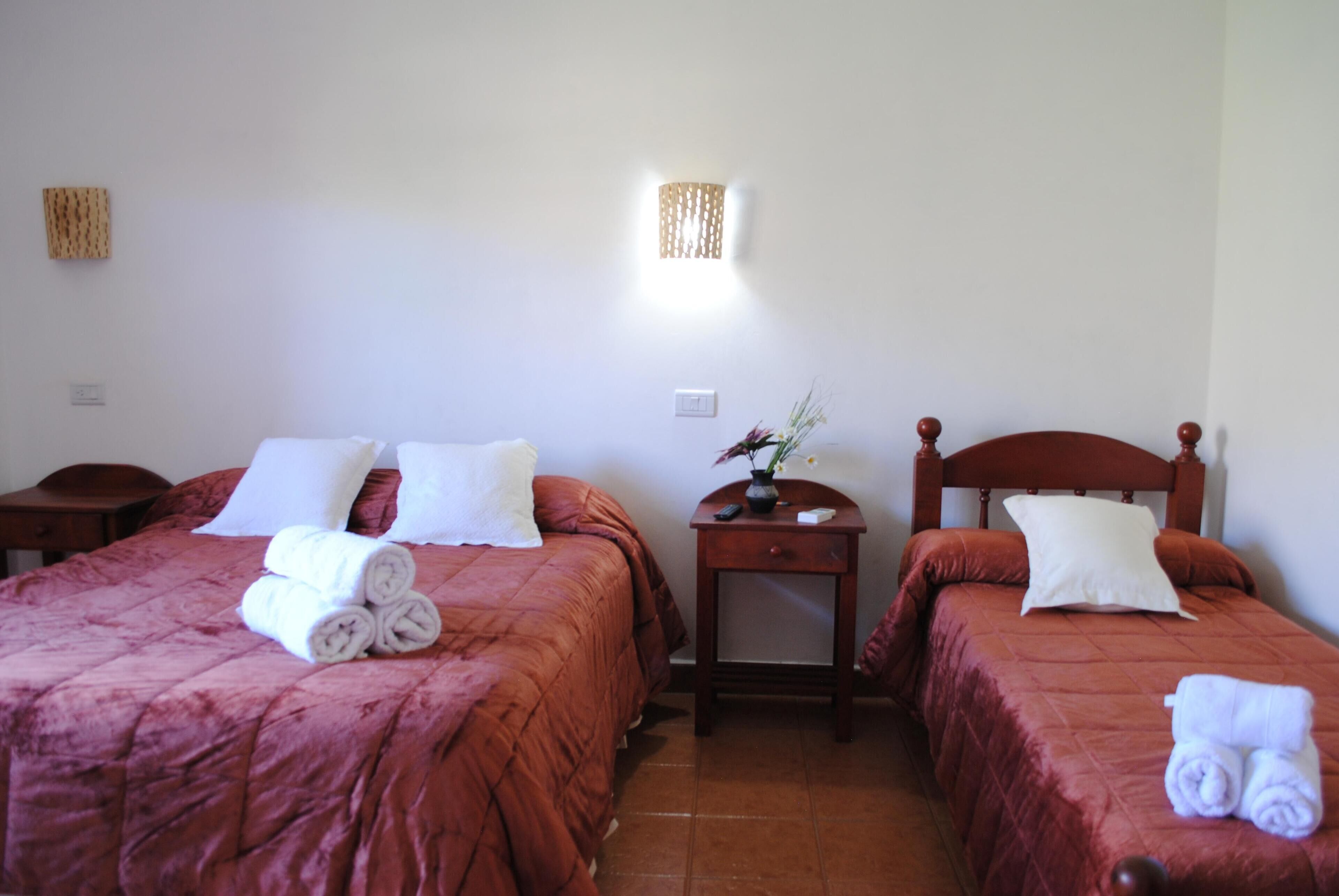A hotel room with two beds, each with a red bedspread and white pillows. There are towels on the beds and a small table with a vase of flowers between the beds.