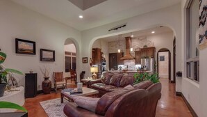A spacious living room with a leather sofa, a coffee table, and a dining area with chairs. There is a kitchen visible in the background with modern appliances and a stone backsplash. - Resort Style S/W Home on 3 Acres. Views, Pool, Outdoor Fireplace (Bulverde)