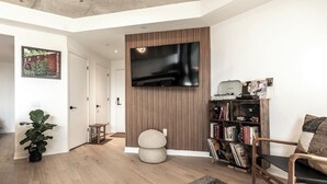 Una sala de estar moderna con un televisor de pantalla plana montado en un panel de madera, una estantería con libros y objetos decorativos, un sillón cómodo y una planta grande en maceta.