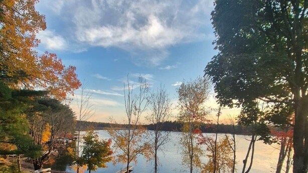 A stone patio with Adirondack chairs and a fire pit, overlooking a lake with a dock and boats. - Waterfront 3-Bedroom Cottage with Hot Tub, Fire Pit, Kayaks (Ontario)