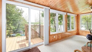 A room with large sliding glass doors leading to a wooden deck, a ceiling fan, and a view of a wooded area. - Waterfront 3-Bedroom Cottage with Hot Tub, Fire Pit, Kayaks (Ontario)