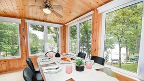 A dining area with a wooden table set for four, surrounded by large windows offering a view of a wooded area. - Waterfront 3-Bedroom Cottage with Hot Tub, Fire Pit, Kayaks (Ontario)