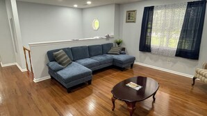 Ein Wohnzimmer mit Ecksofa, rundem Couchtisch, Holzboden, Fenster mit Blick und Einbauleuchten.