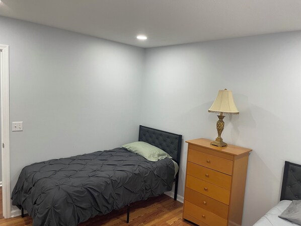 A small, neatly arranged bedroom with a single bed, a dresser, and a bedside lamp.