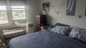 Ein Schlafzimmer mit Bett, Kommode, Schreibtisch, Lampe und Fenster mit Blick auf Häuser und ein Gewässer.