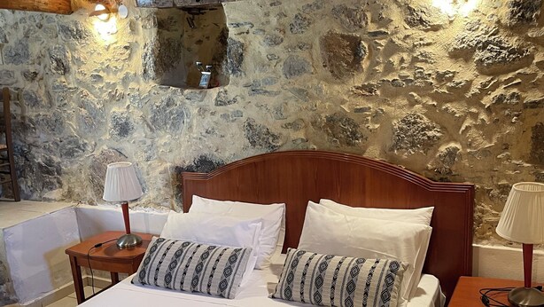 Ein Schlafzimmer mit Steinwand, Holzbalken an der Decke, einem Bettgestell aus Holz und einem Nachttisch mit Lampe.