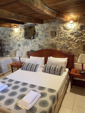 A bedroom with a stone wall, wooden ceiling beams, a wooden bed frame, and a nightstand with a lamp.