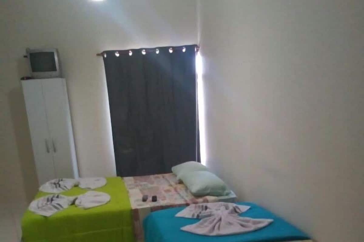 Une petite pièce bien éclairée, avec deux lits, un pouf vert, une armoire et un ventilateur de plafond.