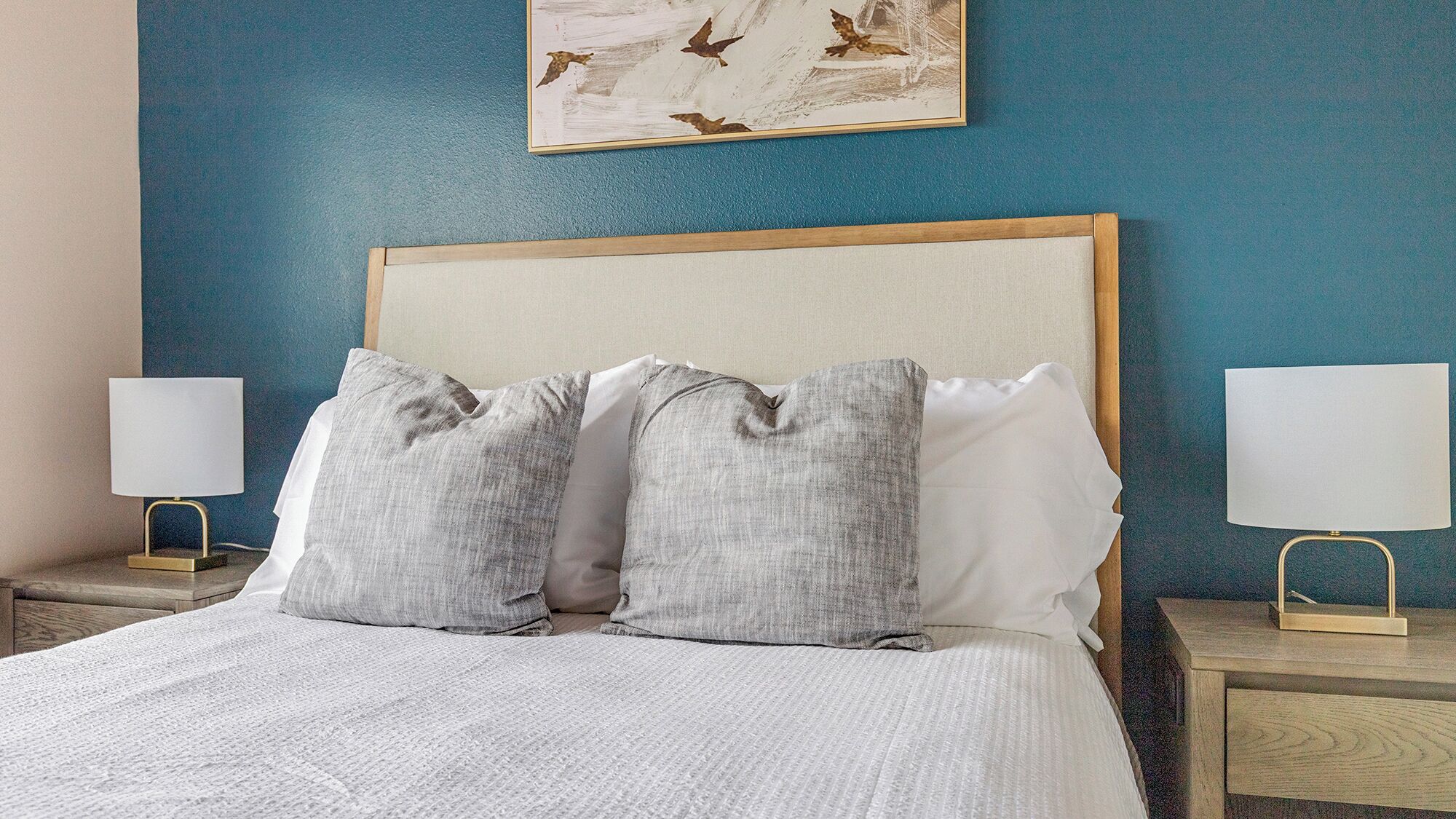 Una cama bien hecha con una colcha blanca y dos almohadas grises, flanqueada por dos mesitas de noche con lámparas, frente a una pared azul con una obra de arte enmarcada.