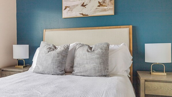 A neatly made bed with a white comforter and two gray pillows, flanked by two bedside tables with lamps, against a blue wall with a framed artwork above. - Landing New Braunfels (New Braunfels)
