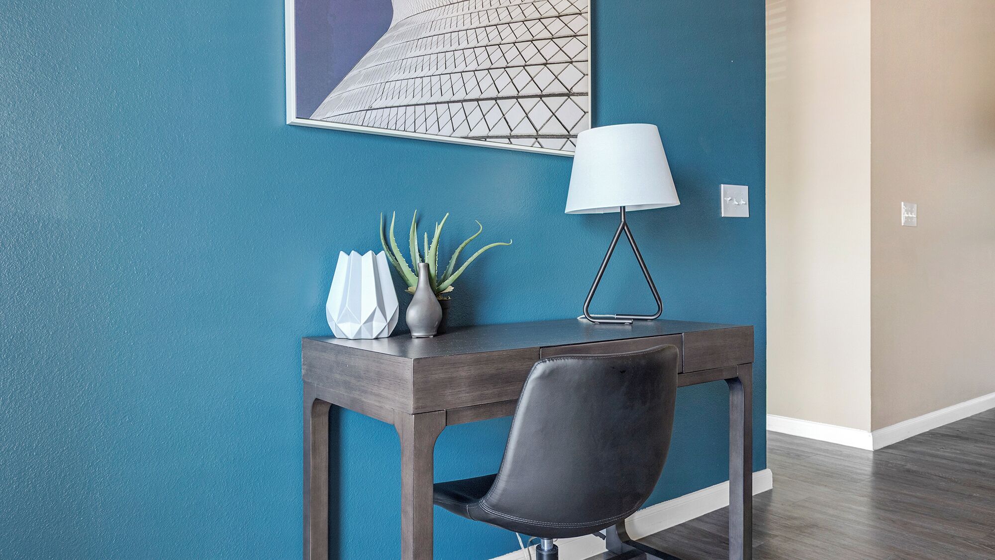 Ein Zimmer mit türkisfarbenen Wänden, einem dunklen Holztisch, einem schwarzen Ledersessel, einer weißen Vase mit Pflanzen und einer modernen Lampe.