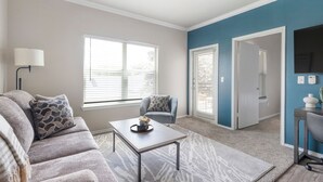 A living room with a sofa, a coffee table, and a chair. There is a window with blinds, a door leading to another room, and a wall-mounted TV. - Landing Furnished Apartments Westover Hills (San Antonio)