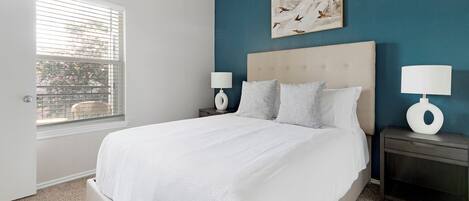 Een slaapkamer met een groot bed, twee nachtkastjes met lampen, een raam met zonwering en een muur met een schilderij.