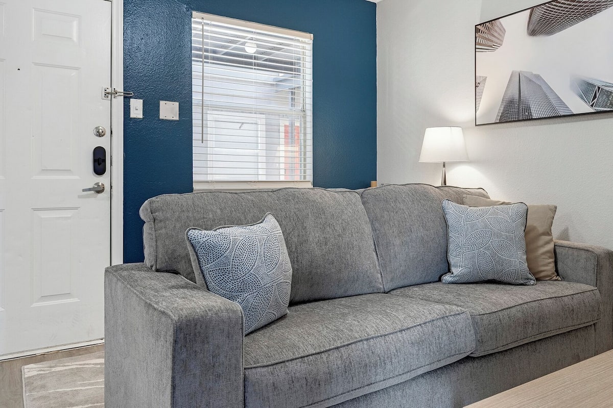 A grey sofa with patterned cushions, a white door, a window with blinds, a lamp, and a framed picture on the wall.