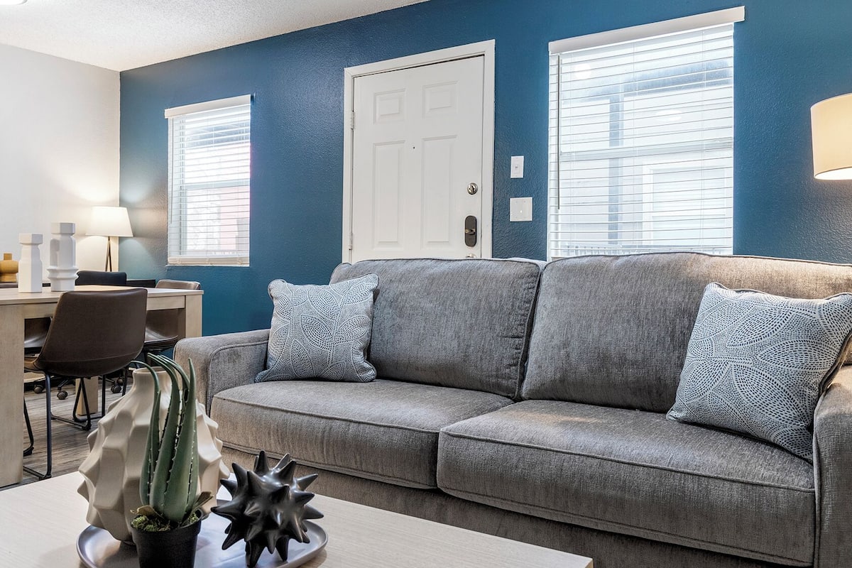 A living room with a gray sofa, two patterned pillows, a cactus, and a star-shaped decoration on a coffee table. There is a dining area with a table and chairs, and a lamp on a side table.