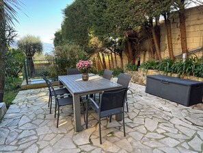 An outdoor patio with a stone floor, a wooden table with chairs, a black sofa, and a flower vase.