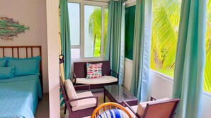 A room with a bed, a sofa, a dining table, and chairs. There are windows with curtains, a ceiling fan, and a view of palm trees outside.