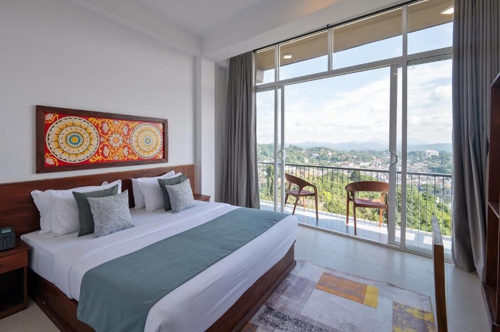 A modern hotel room with a large bed, a view of the city through floor-to-ceiling windows, and a balcony with chairs.