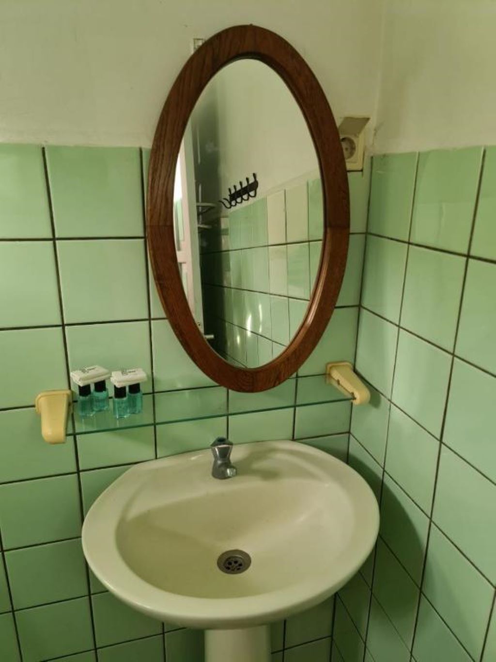 Un baño con pared de azulejos verdes, un lavamanos blanco, un espejo con marco de madera y dos dispensadores de jabón.