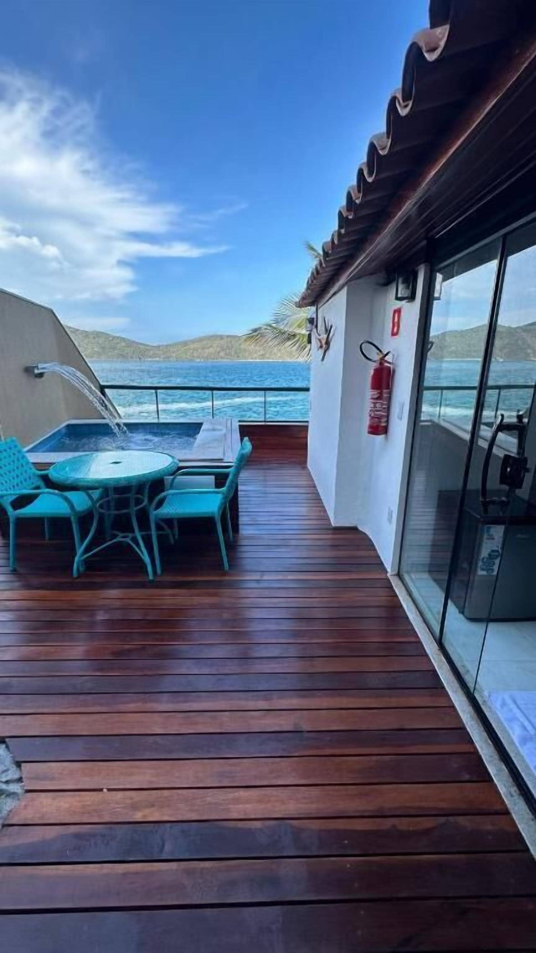 Una terraza de madera con una mesa y sillas, un extintor y una pared de vidrio con vistas al océano.