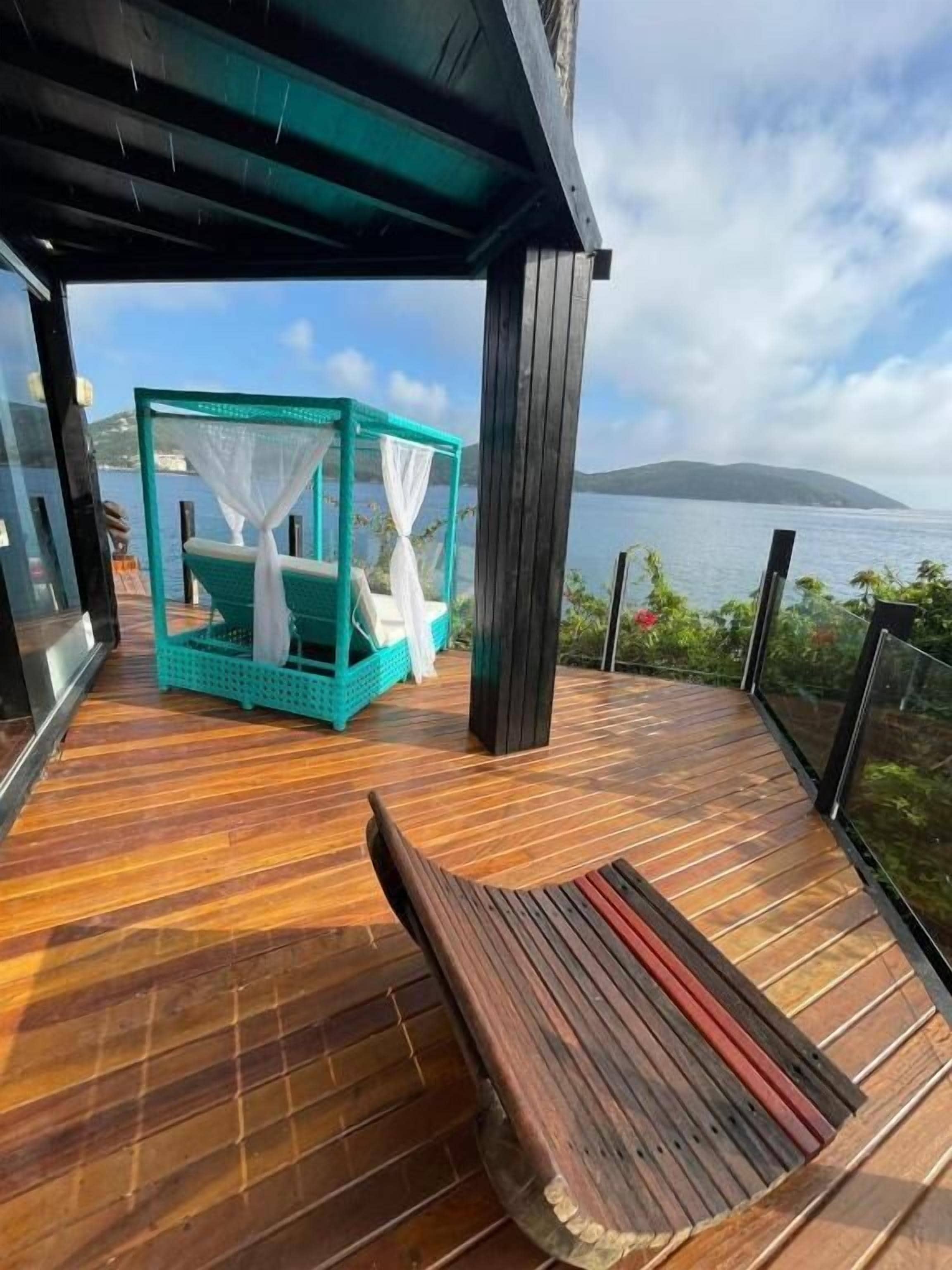 A wooden deck with a bench, a turquoise daybed with white curtains, and a view of the sea and a distant island.