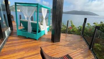 A wooden deck with a bench, a turquoise daybed with white curtains, and a view of the sea and a distant island.