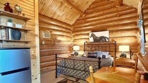 A log cabin interior with a bed, a wooden dining table, a refrigerator, and a ceiling fan.