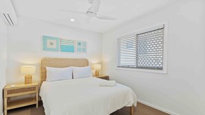 A hotel room with a bed, two bedside tables with lamps, a ceiling fan, a window with blinds, and wall art. - Thirty-Nine Albatross (Apartment 8) (Gold Coast)