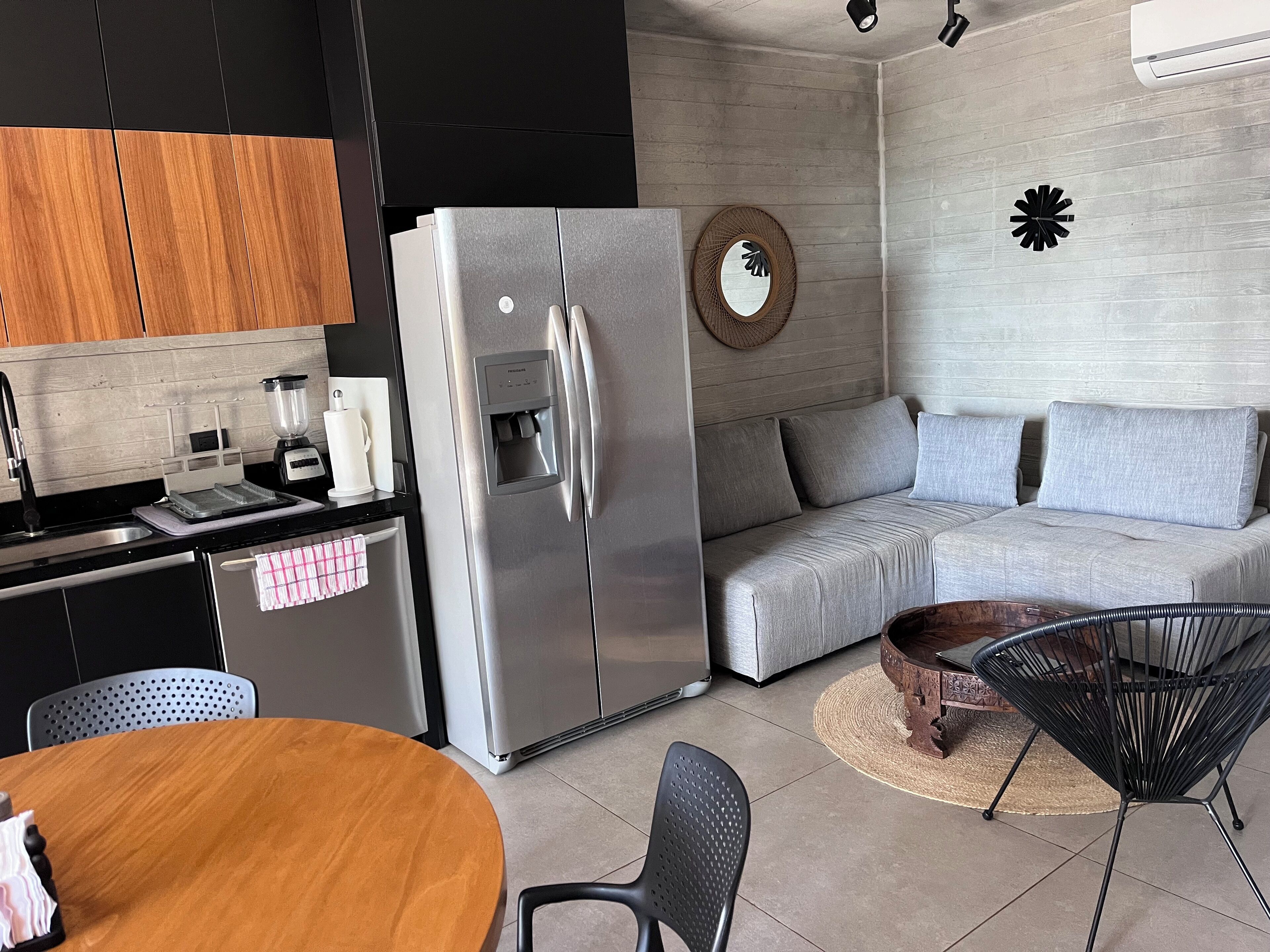 Una cocina moderna con un refrigerador de acero inoxidable, una cafetera y un lavabo. Hay un sofá gris y una mesa de comedor de madera con sillas.