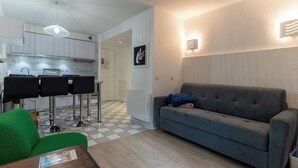 Ein stilvoller Wohnbereich mit einem grauen Sofa, einem grünen Sessel und einem kleinen Tisch. Im Hintergrund befindet sich eine Küche mit weißen Schränken und einem gefliesten Boden.