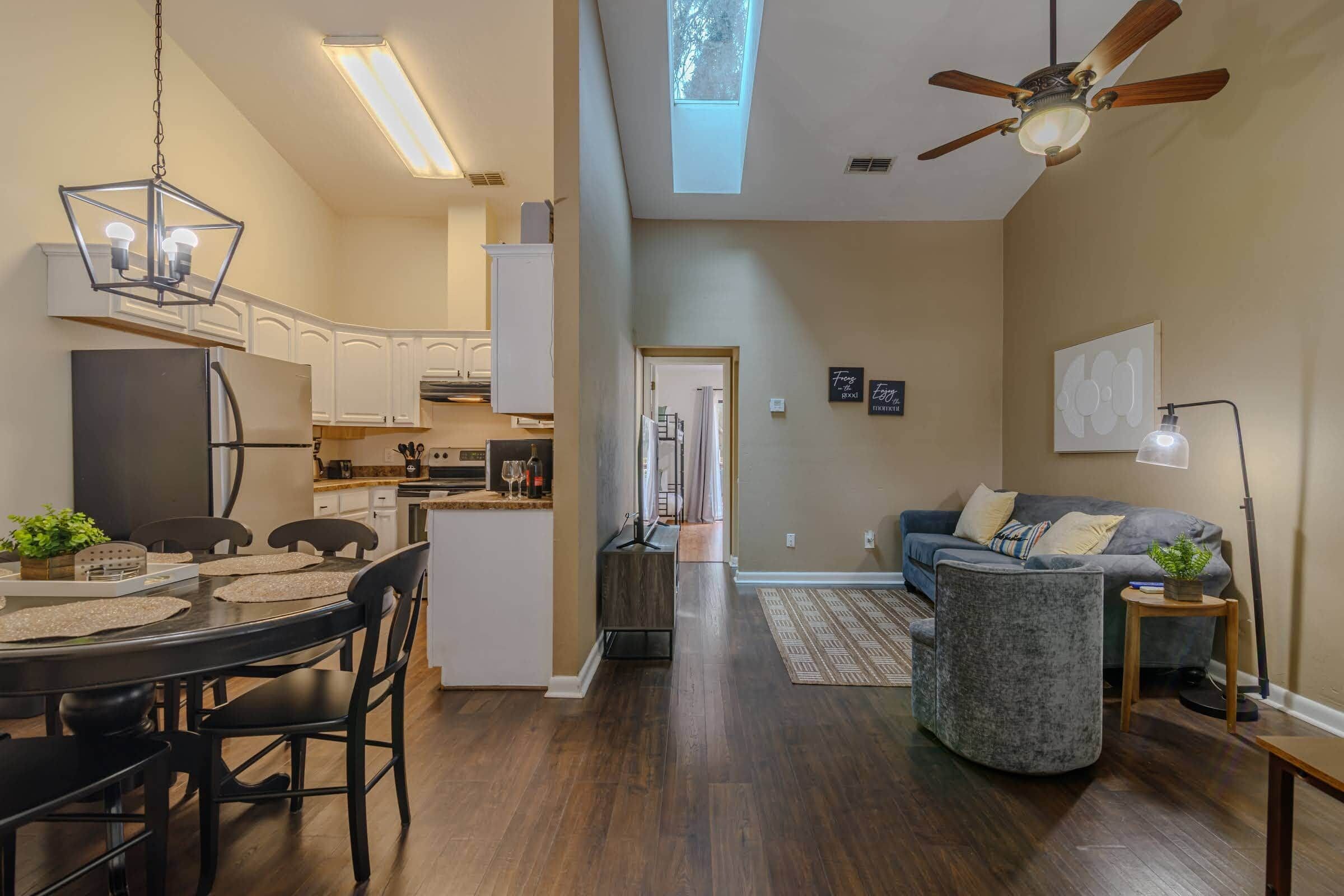 A spacious living area with a dining table, kitchen in the background, and a ceiling fan.