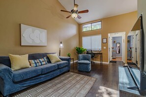 A living room with a blue sofa, a patterned rug, a ceiling fan, and a window with blinds.