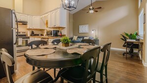 A modern kitchen with white cabinets, a dining area with a round table and chairs, a ceiling fan, and a living area with a sofa and a plant. - Ultimate Getaway: Yard, BBQ & Games (Gainesville)