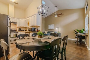 A modern kitchen with white cabinets, a dining area with a round table and chairs, a ceiling fan, and a living area with a sofa and a plant.