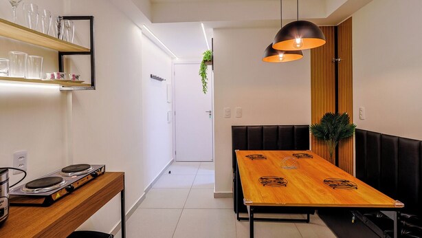 Una cocina moderna con una mesa de madera y taburetes negros, una lámpara colgante y una estantería con vasos.